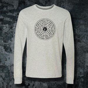 May include: A light gray long-sleeve shirt featuring a circular graphic with text and a star design in the center. The text includes phrases like "Closer to the Heart" and "Fly by Night". The shirt is made of a soft material.