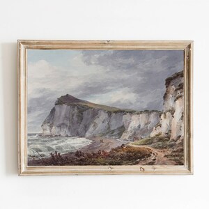 May include: A framed watercolour painting depicting a coastal scene with white cliffs, a rocky beach, and a cloudy sky. The painting is framed in a light wood frame.