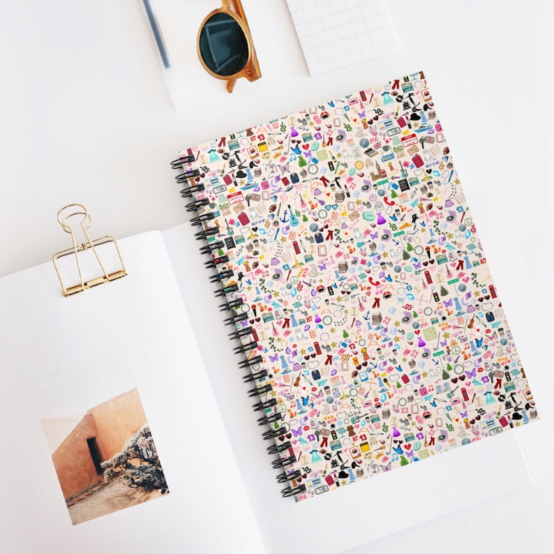 May include: Spiral-bound notebook with a cream-colored cover densely covered in colorful, tiny illustrations. The illustrations include clothing, musical notes, and other small objects. The notebook is open on a white surface, with a gold paper clip and sunglasses nearby.