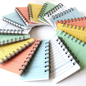 May include: A set of 10 colorful spiral bound notebooks with lined pages. The notebooks are arranged in a circle with the spines facing outwards. The notebooks are all different colors, including green, yellow, orange, pink, and blue.