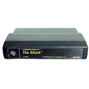 May include: Black Command Module video game cartridge labeled "The Attack*" with text and a Texas logo. The label includes "COMMAND MODULE", "1980 TI", and "TRADEMARK OF MILTON BRADLEY CO".