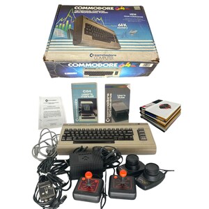 May include: A Commodore 64 personal computer set including the original cardboard box, a beige keyboard unit, two black joysticks with red buttons, a power supply, cables, a user manual, a disk drive guide, and a stack of floppy disks.