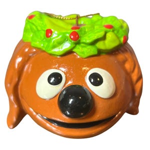 May include: A ceramic ornament shaped like a cartoon dog's head. The dog has brown fur, large white eyes with black pupils, and a black nose. A green wreath with red berries sits atop the head. The ornament is likely for Christmas.