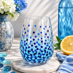 May include: A stemless wine glass with a pattern of blue polka dots. The glass sits on a white marble coaster. A blue vase with white and blue flowers is in the background, along with a blue bottle and a lemon.