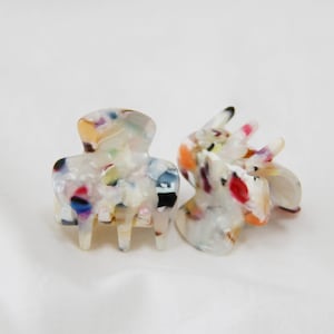 May include: Two small hair claw clips with a marbled effect. The clips feature a mix of white, black, blue, red, and orange colours. They are made from a glossy material and are designed to secure hair.