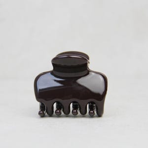 May include: A small, dark brown hair claw clip. The clip is made of glossy plastic and has a rounded top with a series of teeth along the bottom edge. The clip is designed to hold hair in place.