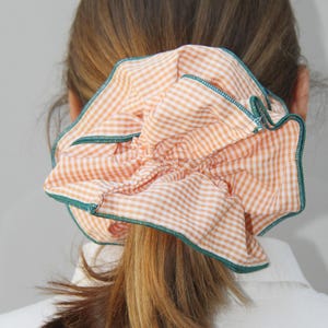 May include: A close-up of a hair scrunchie made of orange and white gingham fabric with a green trim.