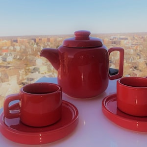 Puede incluir: Una tetera roja y dos tazas de té rojas con platillos. La tetera tiene un cuerpo redondeado y una tapa con un pomo. Las tazas de té tienen una forma redondeada y un asa. Los platillos son redondos y tienen un borde ligeramente elevado.