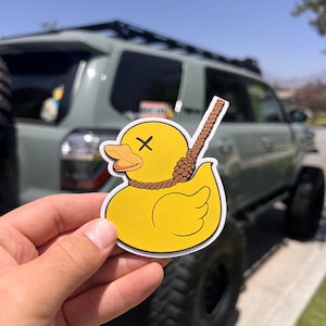 May include: A yellow rubber duck with a brown rope tied around its neck, forming a noose. The duck has an X drawn on its face. The duck is held in a hand in front of a green SUV.