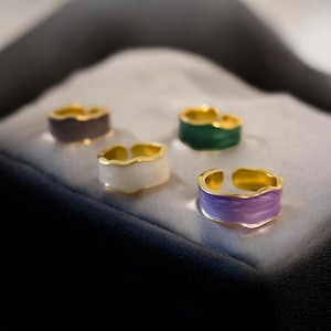 May include: Five gold rings with different colored enamel. The rings are on a black velvet surface. The colors of the enamel are purple, white, green, and gray.