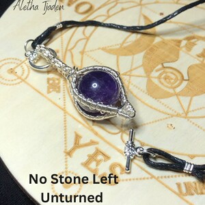 May include: A silver pendant necklace featuring a wire-wrapped design holding a deep purple gemstone. The necklace has a black cord and a toggle clasp. The text "No Stone Left Unturned" is at the bottom of the image.