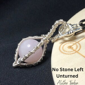 May include: A silver wire-wrapped pendant featuring a light pink sphere. The pendant has an intricate cage-like design and is suspended from a black cord. The text "No Stone Left Unturned" is visible in the image.