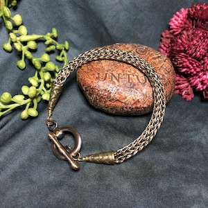 May include: A silver-coloured bracelet with a woven chain design. The bracelet features an ornate toggle clasp and decorative end pieces. The jewellery is displayed on a textured stone and a dark blue surface.