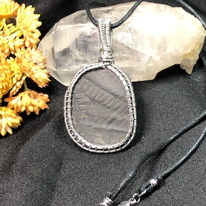 May include: A wire-wrapped pendant necklace featuring a dark gray, oval-shaped stone with a textured surface. The pendant is suspended from a black cord with a silver-toned clasp. The necklace is displayed against a dark background.