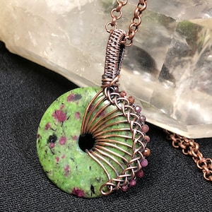 May include: A handmade pendant necklace featuring a green stone with red inclusions, wire-wrapped in copper. The pendant is donut-shaped with a central hole. The necklace has a copper chain and is displayed against a neutral background.