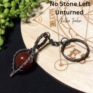 May include: A handcrafted keychain featuring a reddish-brown gemstone encased in a copper-colored wire wrap. The keychain includes a hammered metal ring and a small chain. The text "No Stone Left Unturned" and "Aleta Tjaden" are visible.