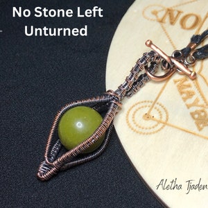 May include: A handcrafted pendant featuring a green stone encased in copper wire. The wire is intricately woven, forming an eye-like shape. The pendant hangs from a black cord with a copper clasp. Text on the image reads "No Stone Left Unturned."