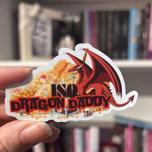 Op de afbeelding: Een sticker met een rode draak met zwarte vleugels, bovenop een stapel gouden munten en schatten. De sticker heeft de tekst "ISO DRAGON DADDY" in een rood en wit lettertype.