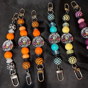 Puede incluir: Seis llaveros de cuentas con cierres plateados. Los llaveros presentan cuentas en naranja, negro, amarillo, turquesa y morado. Cada llavero tiene un dije circular con la palabra "Anxiety" y un diseño de mano de esqueleto.