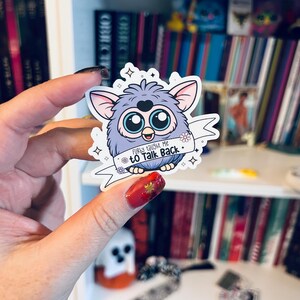 May include: A sticker featuring a cartoon Furby character with blue and purple fur, large blue eyes, and a banner that reads "Furby taught me to talk back." The sticker has a white border and is held up against a bookshelf.