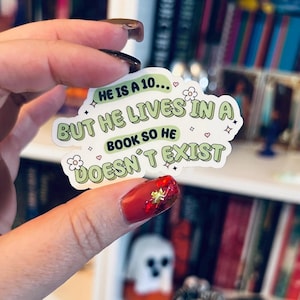 May include: A white sticker with green text that reads "HE IS A 10... BUT HE LIVES IN A BOOK SO HE DOESN'T EXIST." The sticker has a light green border and is decorated with small flowers, hearts, and stars.