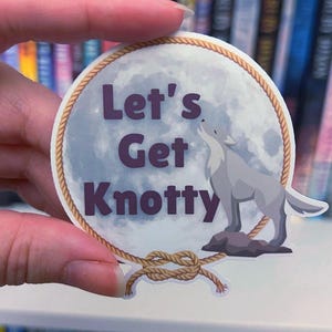 Pode incluir: Um adesivo redondo com uma borda de corda e um fundo de lua cheia. O texto "Let's Get Knotty" está em letras roxas. Um lobo cinza está a uivar para a lua.