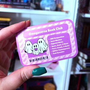 May include: A pink card with the text "Omegaverse Book Club" and a list of topics related to the genre. The card features three white ghosts with hearts on their chests.