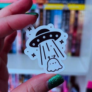 Puede incluir: Una pegatina blanca con una ilustración en blanco y negro de un OVNI que lleva a un pequeño extraterrestre amigable. La pegatina tiene un fondo blanco con estrellas negras y rayos de luz.
