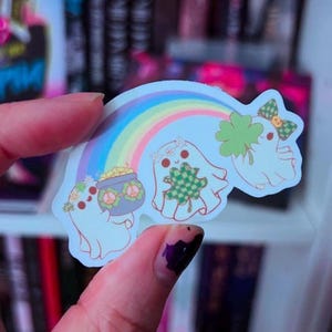 May include: A colorful sticker featuring three cartoon ghosts with St. Patrick's Day and rainbow themes. One ghost holds a pot of gold, another a shamrock, and the third has a shamrock bow. The sticker has a rainbow arching over the ghosts.
