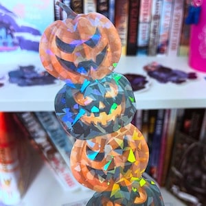 May include: A stack of four iridescent holographic pumpkin stickers with jack-o-lantern faces. The stickers are stacked on top of each other and are in a hand.