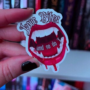 May include: A sticker with a red mouth design, featuring sharp fangs and dripping blood. The sticker has the words "Love Bites" and "...and so do I" in a gothic font. The sticker is white with a red and black design.