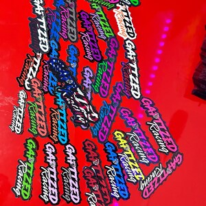 Puede incluir: Una colección de pegatinas coloridas con el texto "GATIZED Racing" en diferentes fuentes y colores. Las pegatinas están dispuestas en un patrón aleatorio sobre una superficie roja.