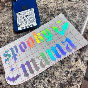Puede incluir: Un teléfono inteligente azul con una pegatina de código QR y el texto "FYE.DESIGNS1" junto a una calcomanía de vinilo holográfico. La calcomanía presenta la palabra "Spooky Mina" en una fuente gótica, junto con formas de estrella y murciélago.