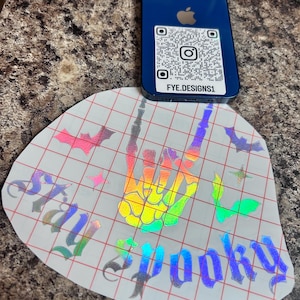 May include: A blue smartphone rests on a sticker with a rainbow hand gesture and the words "Stay Spooky." The sticker also features bats and stars. The phone has a QR code and the text "FYE.DESIGNS1."