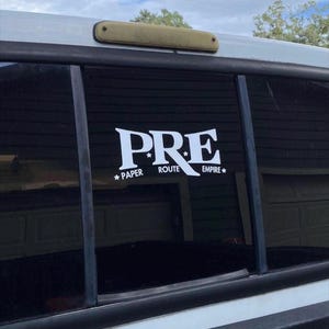 May include: A white decal on a vehicle window features the text "PRE" in a stylized font, with "PAPER ROUTE EMPIRE" below. The decal is affixed to a black-tinted window, with a light-colored vehicle body visible.