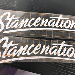 Puede incluir: Dos calcomanías de vinilo blanco con la palabra "Stancenation" en fuente cursiva. Las calcomanías están sobre un fondo negro con un patrón de cuadrícula. Las calcomanías son rectangulares y tienen un respaldo transparente.