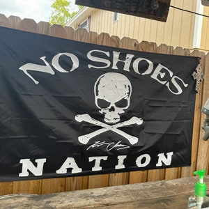 Op de afbeelding: Een zwarte vlag met een wit schedel- en kruisbeenderontwerp. De tekst "NO SHOES NATION" is in wit op de vlag gedrukt.