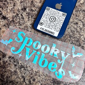 Puede incluir: Un teléfono inteligente azul con un código QR y el identificador de Instagram FYE.DESIGNS1. También es visible una pegatina transparente con las palabras "Spooky Vibes" en escritura turquesa, junto con gráficos de murciélagos y estrellas.