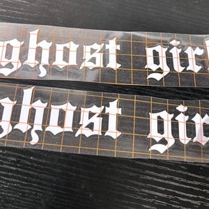 May include: Two white vinyl decals featuring the words "ghost girl" in a gothic font. The decals are on a clear backing with a grid pattern, and are placed on a dark surface.