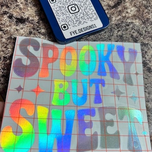Puede incluir: Una pegatina holográfica con las palabras "Spooky But Sweet" en letras de burbujas de color arcoíris. La pegatina está sobre un fondo de cuadrícula gris. Un teléfono azul con un código QR y el texto "FYE.DESIGNS1" está en el fondo.