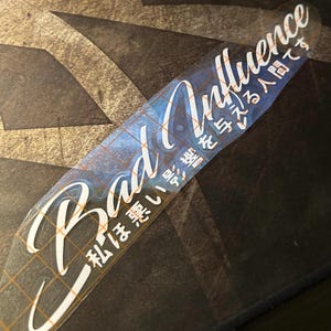 May include: A clear sticker with the words "Bad Influence" in a stylized white font. Japanese characters are also present. The sticker has a grid pattern on the back and is placed on a dark, textured surface.