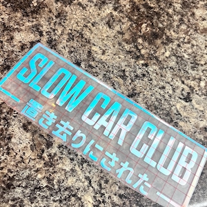 Puede incluir: Una pegatina rectangular con las palabras "SLOW CAR CLUB" en una fuente turquesa brillante. Debajo del texto en inglés hay texto japonés del mismo color. La pegatina está sobre un respaldo transparente.