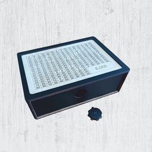 May include: A black wooden box with a grid of numbers and the text "5.000" printed on the front. The box has a small round hole on the top.
