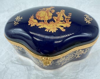 May include: A navy blue, oval-shaped trinket box with gold accents. The lid features a gold illustration of two figures in a garden setting. Gold floral designs adorn the sides and edges of the box, creating an elegant aesthetic.