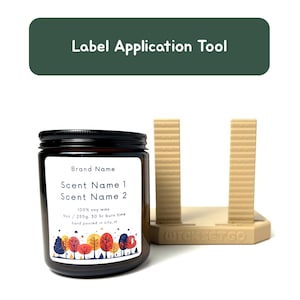 May include: A brown glass jar candle with a black lid and a white label featuring text and a colorful tree design. The label reads "Brand Name", "Scent Name 1", and "Scent Name 2". A beige label application tool is to the right.