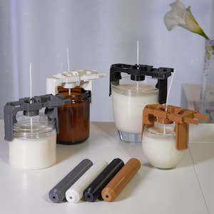 Puede incluir: Se muestran varias herramientas para hacer velas, incluidos dispositivos de centrado de mechas de velas grises, blancas, negras y marrones. Los dispositivos se colocan encima de frascos de vidrio y candelabros llenos de cera. También se muestran varias herramientas de centrado de mechas en varios colores.