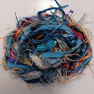 May include: A ball of colorful craft yarn with a mix of textures and colors. The yarn features shades of blue, orange, silver, and red, with a variety of ribbons, threads, and textured fibers. This yarn is suitable for various crafting projects.
