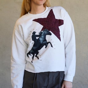 May include: White sweatshirt featuring a large burgundy star and a graphic of a rider on a rearing black horse. The sweatshirt has a crew neck and long sleeves. The design is centered on the front of the shirt.