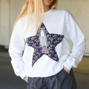 May include: White sweatshirt with a black star print featuring a cartoon figure with pink boots. The star is filled with a floral pattern.