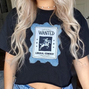 May include: Black cropped t-shirt with a denim patch that says "WANTED LIBERAL COWBOY" with a drawing of a cowboy riding a horse.